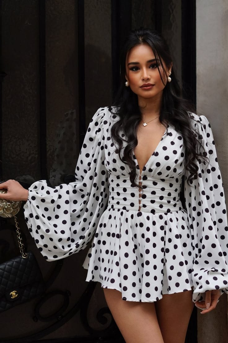 Woman posing in a white polka dot romper dress with dramatic puff sleeves outdoors by a metal gate.