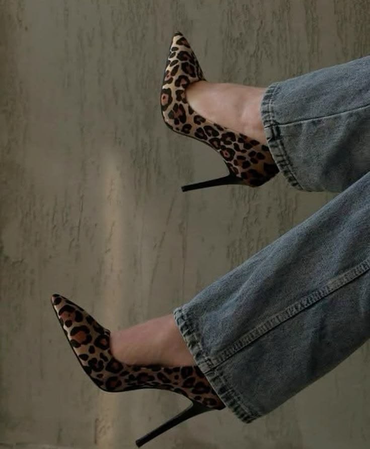 leopard print stiletto heels worn with blue jeans, legs crossed against a neutral wall.
