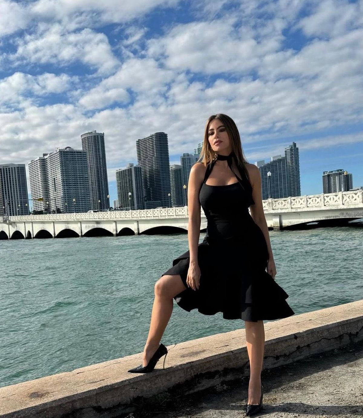 Sofia Vargas' Black Dress and heels posing on a seawall by turquoise water, with a skyline of tall buildings and a white bridge in the background