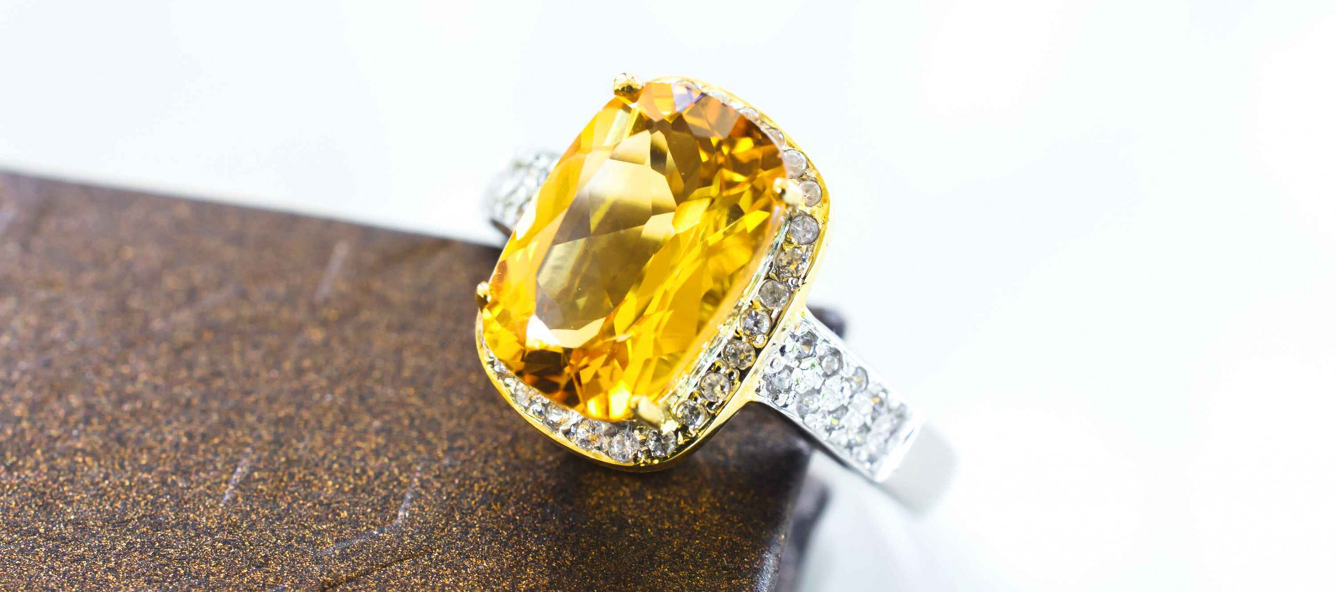 Gold ring with a large yellow sapphire gemstone and diamond halo, resting on a brown textured surface.