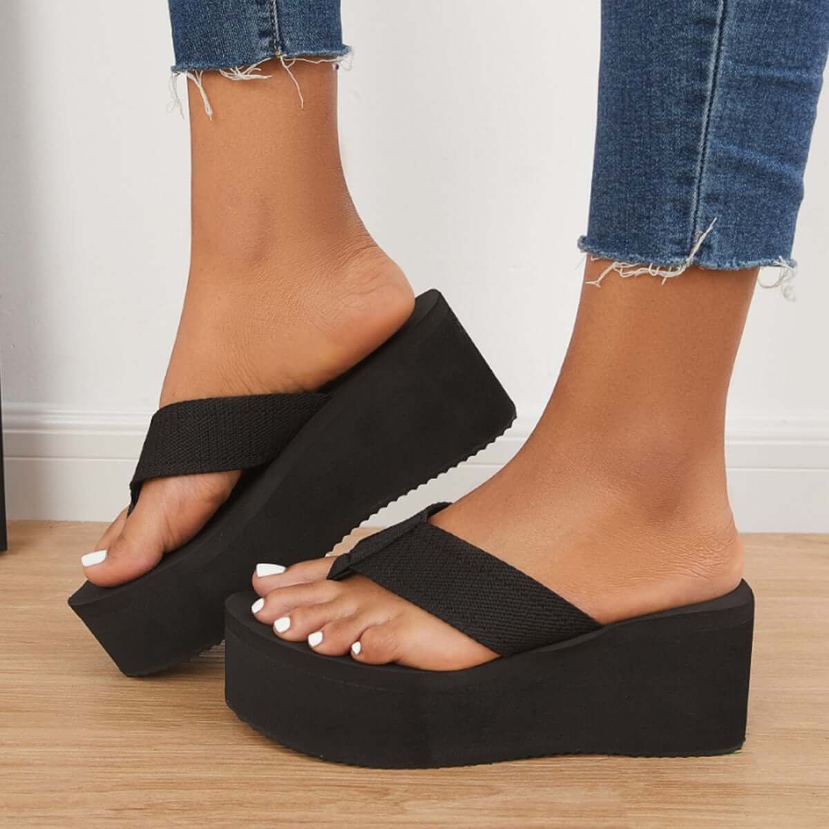 Close-up of a person wearing black platform wedge slides with thick soles and white toenail polish, denim jeans with frayed hems show the ankles.