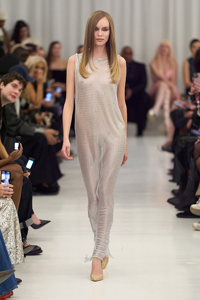 Model walking a runway in a light beige sleeveless jumpsuit with nude heels; audience seated along the catwalk.