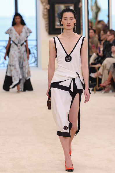 Model walking a runway in a white dress with black trim and a side knot, orange heels, and a small handbag on the left.