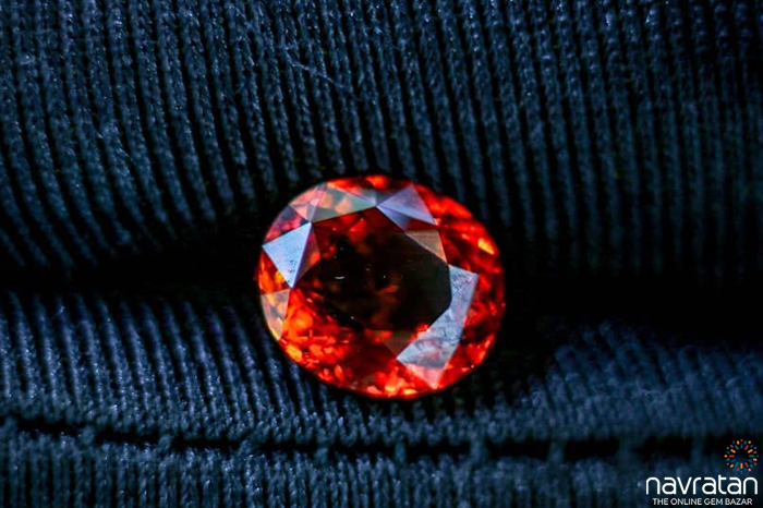 Red faceted gemstone resting on dark blue fabric with a Navaratna logo in the corner