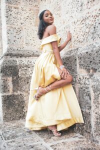 Woman in a yellow off-shoulder gown with a tiara posing against a rough stone wall.