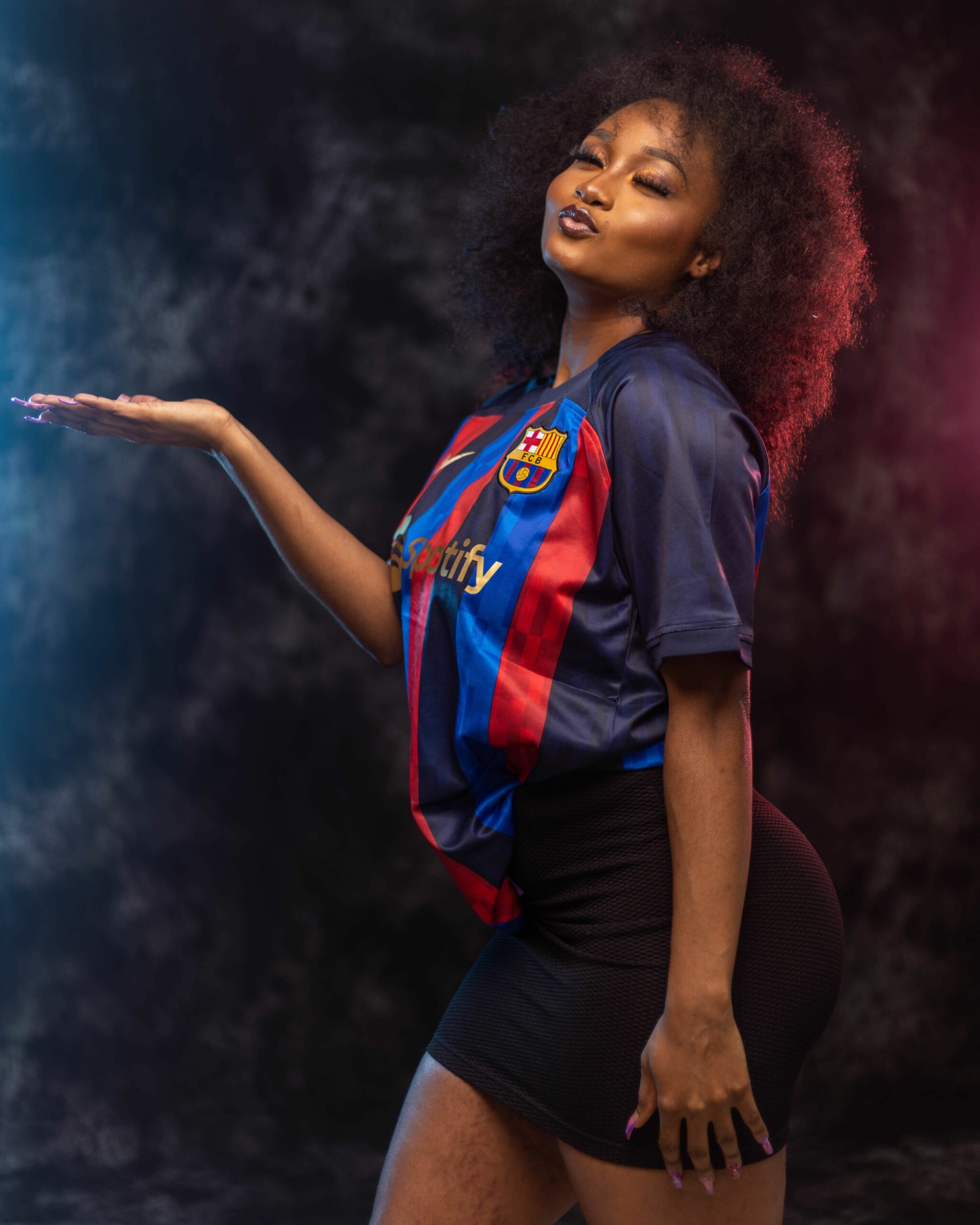 Young woman in a FC Barcelona jerseys poses with an outstretched arm against a blue-pink smoky backdrop.