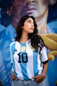 Young woman wearing an Argentina national team jersey (blue and white stripes) with the number 10, looking to the right in front of a colorful mural.