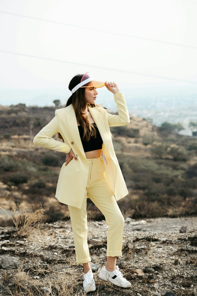Woman in a pale butter yellow pantsuit, black top, and white sneakers stands on a rocky hillside, adjusting an orange visor.