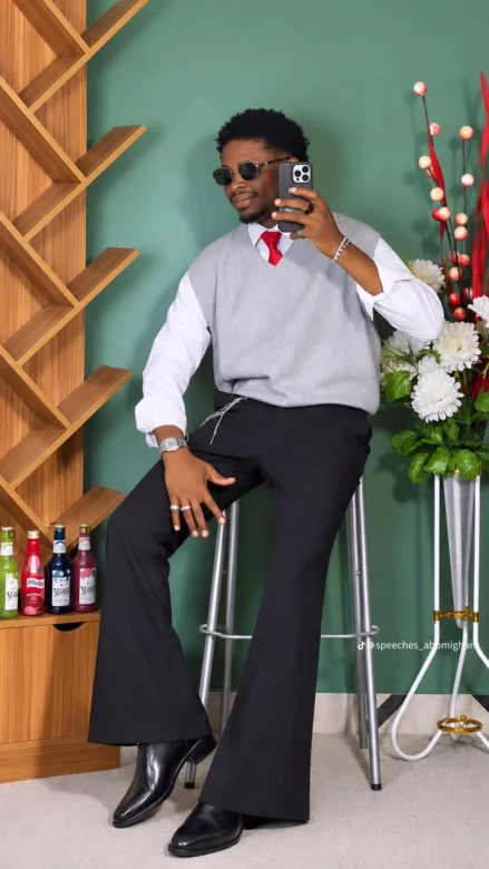 Man in a gray vest, white shirt, red tie, black wide-leg pants taking a selfie with a phone in a green-walled room with flowers and shelves behind him.