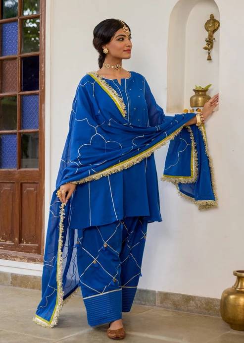 Woman in royal blue traditional Farshi Salwar Suits with gold trim, standing beside a white wall and wooden door in a elegant pose.