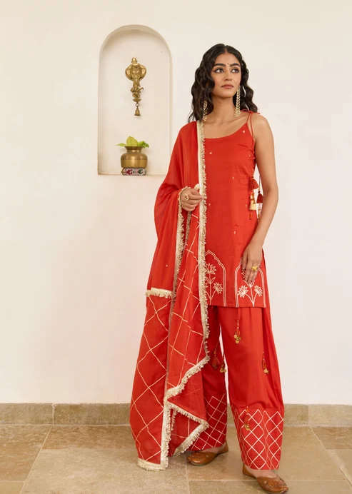 Woman in a red traditional suit with a matching dupatta, standing indoors against a pale wall with a small niche shelf behind her.