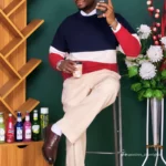 Man in a color-block navy, white, and red sweater poses on a stool, taking a selfie with a smartphone.