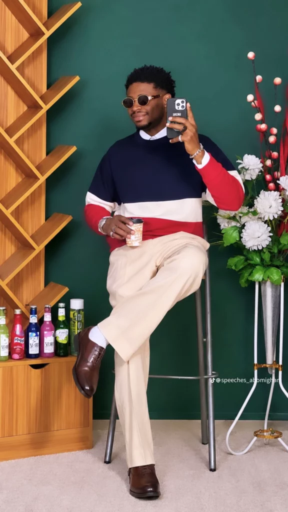 Man in a color-block navy, white, and red sweater poses on a stool, taking a selfie with a smartphone.
