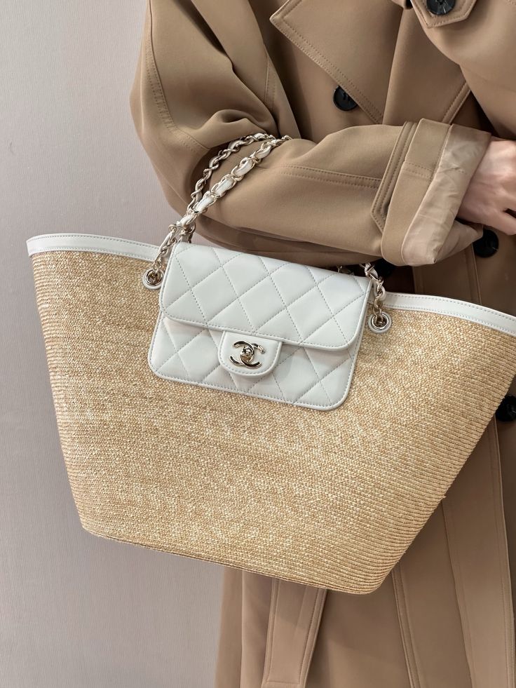 Beige trench coat over a large straw tote bag with a small white quilted purse featuring a Chanel-style clasp hanging from a silver chain strap.