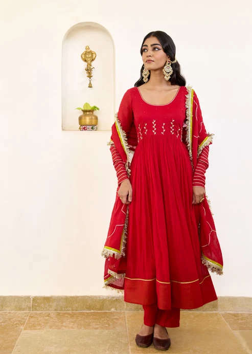 Woman in a bright red traditional Anarkali suit with matching dupatta, standing indoors wearing jhumka earrings.