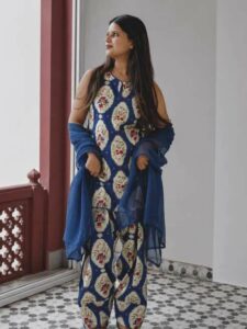 Woman in a blue floral print kurta with matching pants, draped in a sheer blue shawl, standing by a window.