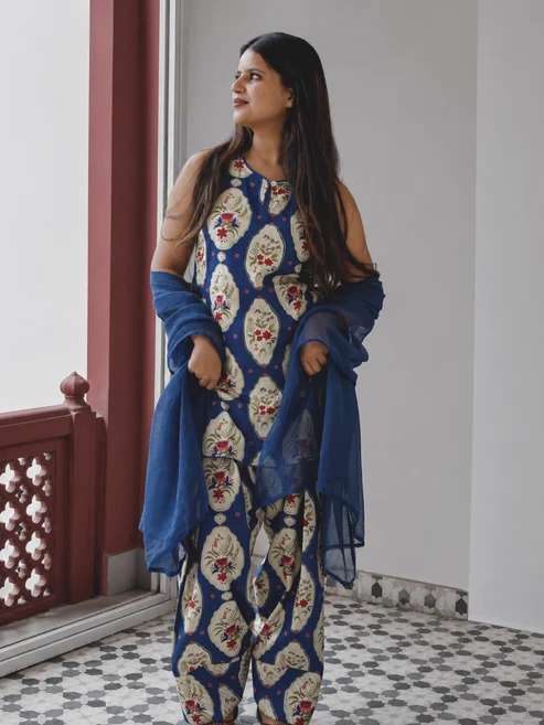 Woman in a blue floral print kurta with matching pants, draped in a sheer blue shawl, standing by a window.