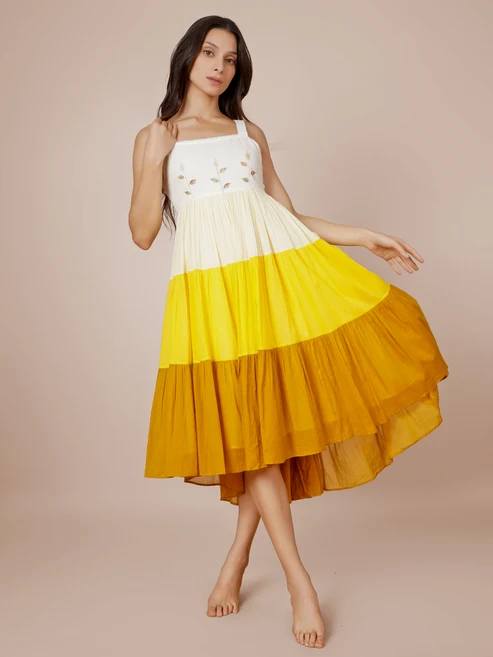 Model wearing a sleeveless color-block, tiered midi dress: white embroidered bodice and gradient yellow to mustard skirt, standing barefoot and posing gracefully.