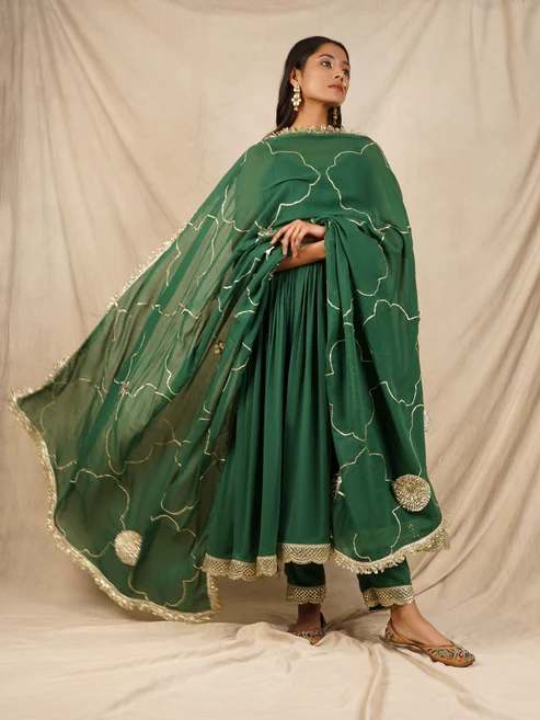 Model wearing a green traditional Indian lehenga with a matching sheer cape and gold-embellished borders, posed against a beige backdrop.