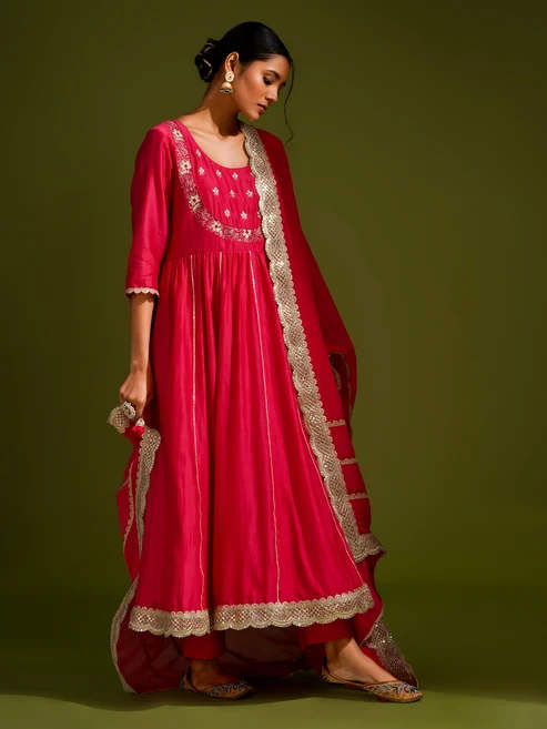 Woman in a bright pink ethnic gown with gold trim, standing sideways against a green backdrop, wearing traditional jewelry and sandals.