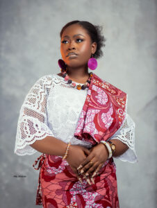 Portrait of a woman in a white lace blouse and bright pink patterned wrap, wearing large pink hoop earrings and a beaded necklace, looking to the left.