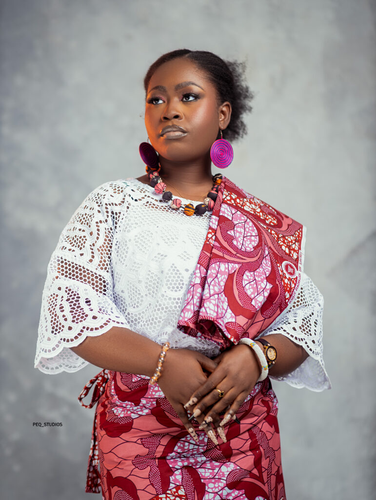 Portrait of a woman in a white lace blouse and bright pink patterned wrap, wearing large pink hoop earrings and a beaded necklace, looking to the left.