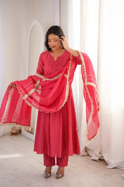 Woman in a pink traditional dress unfurls a matching dupatta with gold trim, standing by a sunlit window.