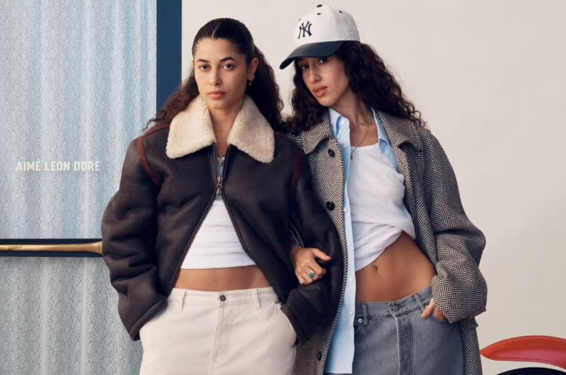 Two women pose in streetwear: one in a brown shearling bomber and white crop top, the other in a checked coat, blue shirt, and a New York Yankees cap.