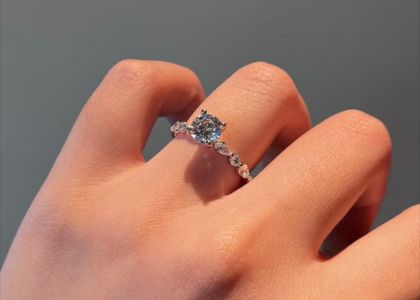Close-up of a hand wearing a diamond engagement ring on the ring finger, featuring a central round diamond with smaller stones along the band.