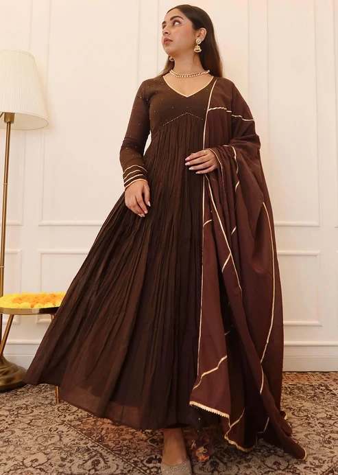 Woman in a brown, floor-length, pleated gown with a matching draped scarf, wearing gold jewelry, standing indoors.