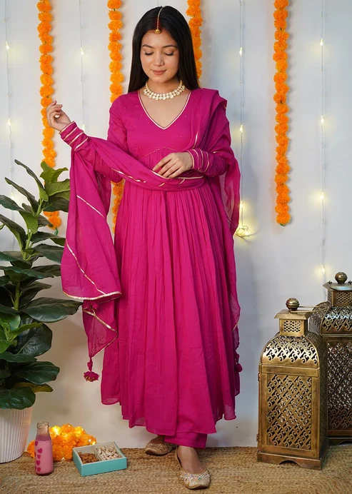 Young woman in a magenta Anarkali suit for women Indian outfit with dupatta, standing indoors against marigold garlands and fairy lights.