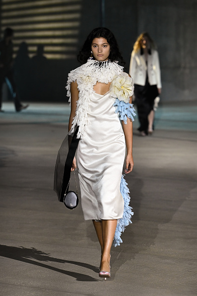 Female model walks a runway in a white satin sheath dress with a large white feather collar and blue feather trim, carrying a black handbag.