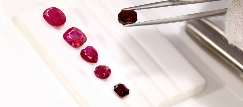 Assorted ruby stone on a white display surface, with metal tweezers holding one gem above them.