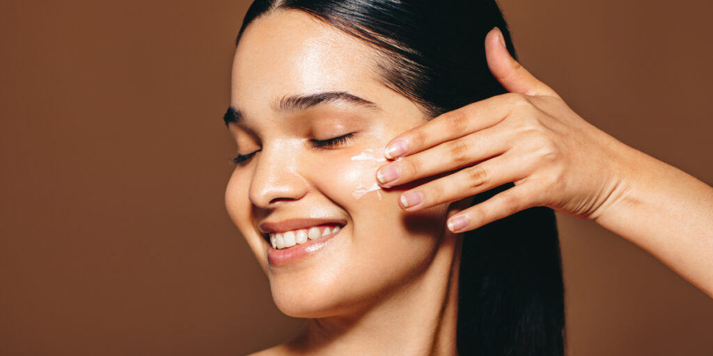 Smiling woman with eyes closed applies cream to her cheek using her fingers during skincare routine.
