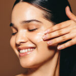Smiling woman with eyes closed applies cream to her cheek using her fingers during skincare routine.