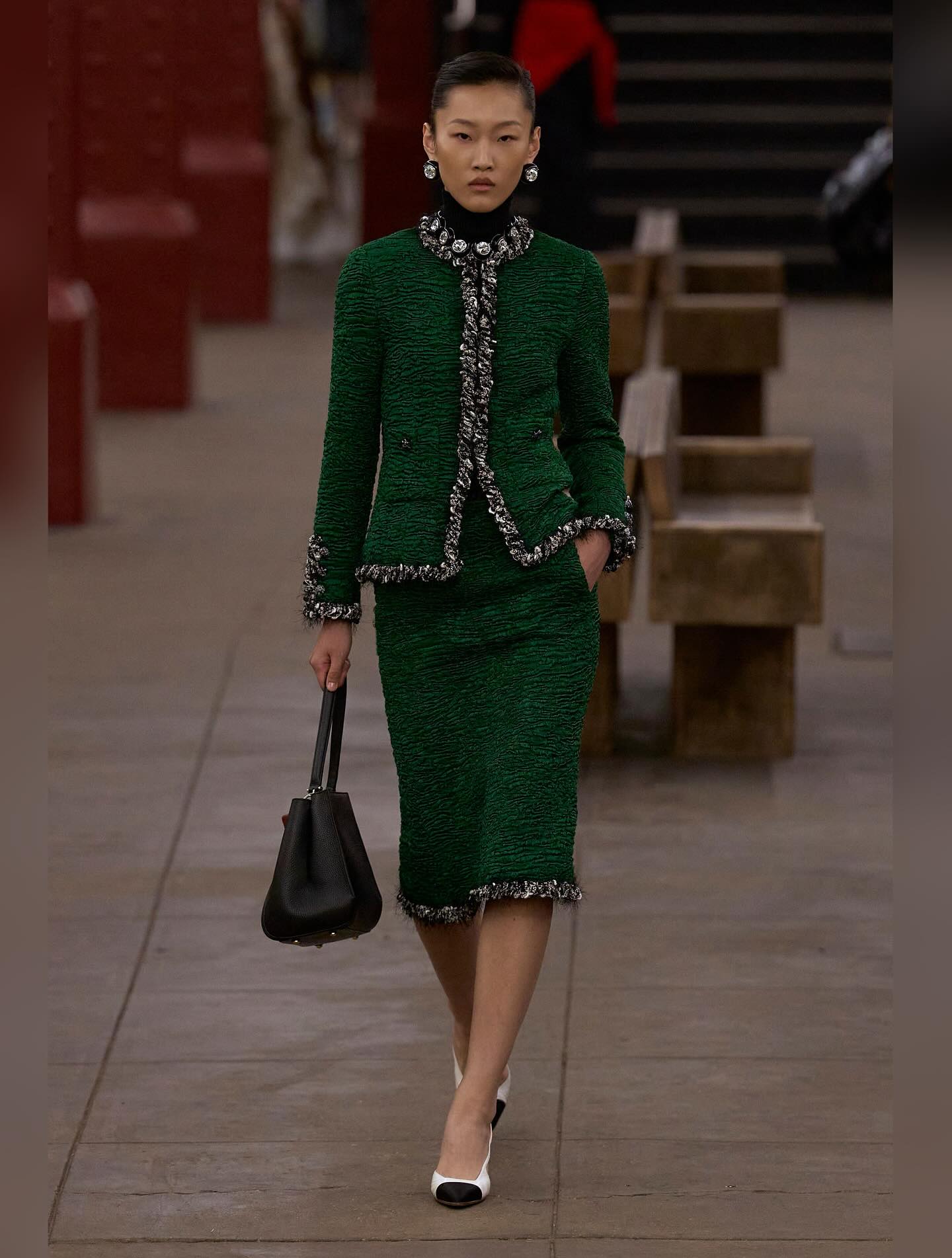 Model Feng Jiao Long walks the runway at Chanel’s Métiers d’Art 2026 ready-to-wear show in an embellished emerald-green silk-tweed jacket and coordinating midi skirt, complemented by two-tone goatskin pumps with mixed-fiber cap toes.