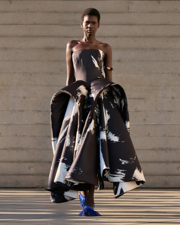 A fashion model strides down the runway at Roksanda’s Spring/Summer 2027 ready-to-wear show in a two-tone black satin midi dress with an off-the-shoulder neckline, sculpted corset bodice, exaggerated peplum, pleated detailing, and white brushstroke accents—a design previously worn by actress Naomi Watts at the 21st Rome Film Festival.
