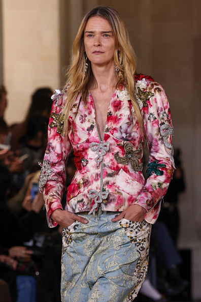 stonian fashion model Carmen Kass appeared on the runway at Andreas Kronthaler’s Vivienne Westwood Womenswear Spring/Summer 2026 show in a long-sleeved ivory blazer adorned with a dense, watercolor-inspired botanical print in vivid fuchsia and deep red, accented by jewel-encrusted brooches, floral fabric rosettes, and dimensional glittering metallic bows studded with rhinestones