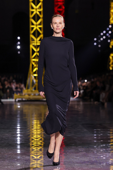 Fashion model Romina Lanaro walks the runway at Chanel’s Womenswear Fall/Winter 2026–2027 show in a long-sleeved black midi dress with a high boat neckline and scooped open back, embellished with a black silk-petal camellia motif evoking delicate feathers