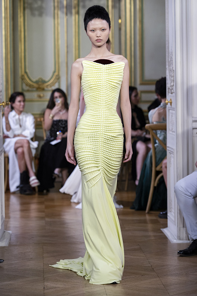 Michael Stewart’s Standing Ground Haute Couture Fall/Winter 2026–2027 collection presented an embellished pale-yellow, form-fitting corseted mermaid gown with a crumb-catcher neckline and a central half-moon panel in black fabric