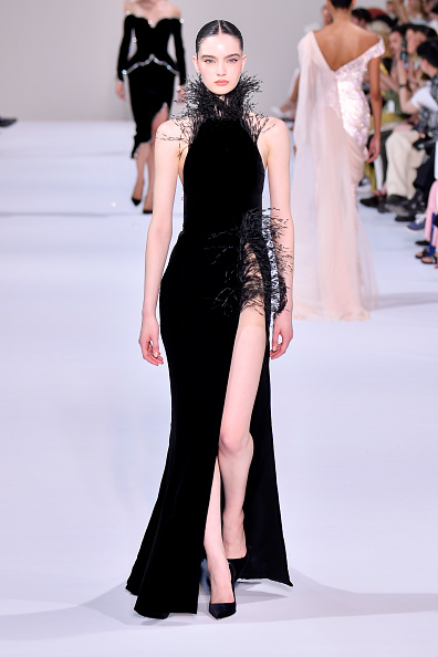 Model Vira Boshkova walked the Tamara Ralph Haute Couture Fall/Winter 2026–2027 runway in a black backless velvet halterneck gown detailed with black dévoré ostrich feathers at the neckline and left hip, a silver crystal-and-white-pearl chain draped across the back, and a high slit rising from the feathered accent