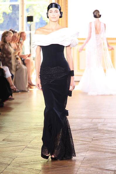 The Georges Chakra Haute Couture Fall/Winter 2026/2027 collection presented an off-the-shoulder black crepe column gown, distinguished by a portrait neckline sculpted in draped ivory silk gazar, an asymmetric beaded-lace panel at the hip finished with a bow, and a lace-edged side slit