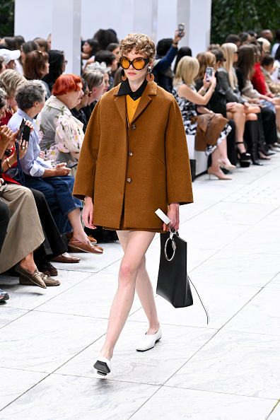 A fashion model walks the runway at the Michael Kors Spring/Summer 2027 ready-to-wear show during New York Fashion Week, wearing a cinnamon tussah bouclé coat over a sunflower cashmere pullover and black poplin dickey, paired with a matching cinnamon tussah bouclé mini skirt