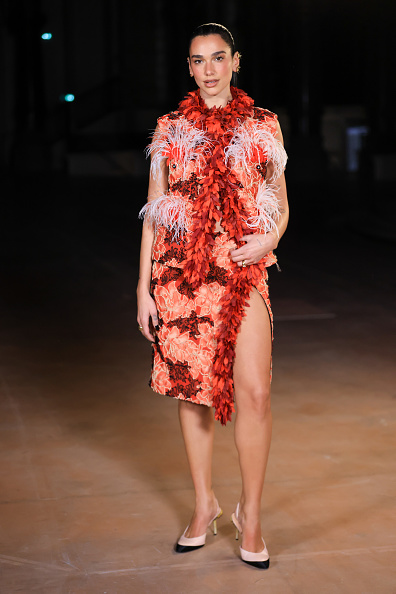 Dua Lipa stole the spotlight at Chanel’s Womenswear Spring/Summer 2027 show in a striking two-tone ensemble, pairing a turtleneck top with a knee-length pencil skirt covered in densely layered coral and cherry-red 3D floral petals and trimmed with a thick, boa-like garland of crimson blooms.