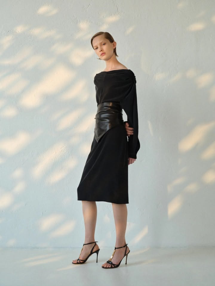 A model wears a black, long-sleeved, knee-length jersey dress from Maximilian Davis’s Ferragamo Pre-Spring/Resort 2027 collection, distinguished by a patent-leather waist corset
